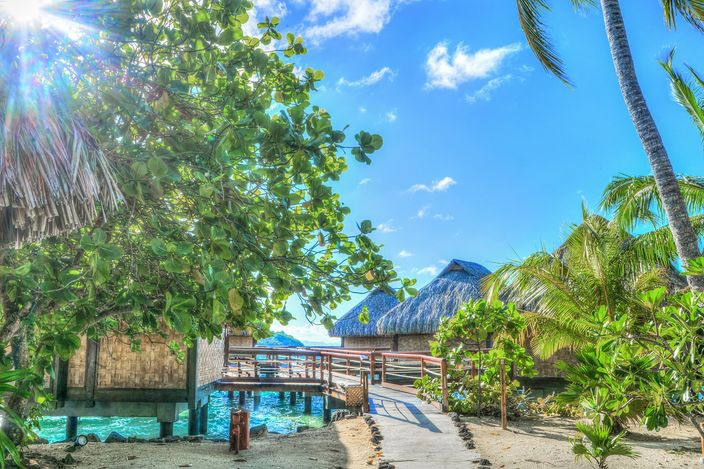 A wooden walkway leading out over a turquoise lagoon inlet to thatched-roof overwater bungalows on stilts, framed by palm and tropical foliage.