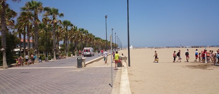A wide, flat sandy beach in Valencia seen from a palm-lined seafront promenade, with people walking and playing on the sand under a clear blue sky, and more beach and buildings stretching into the distance.