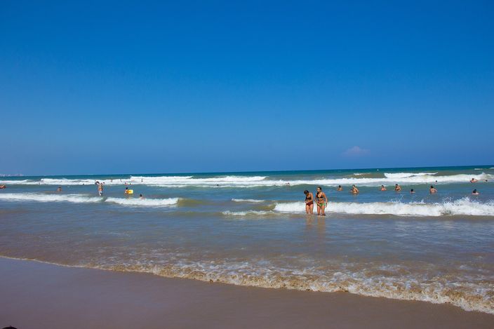 Bathers wading and swimming in small, gentle waves close to shore on a sandy beach, with clear turquoise-blue water and more people visible further along the shoreline under a bright sky.