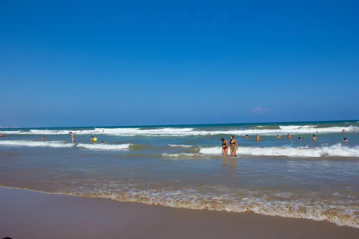 Bathers wading and swimming in small, gentle waves close to shore on a sandy beach, with clear turquoise-blue water and more people visible further along the shoreline under a bright sky.