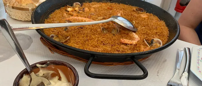 A fideuà in its wide pan on a restaurant table, prawns and clams over toasted noodles, with a clay pot of allioli in the foreground.