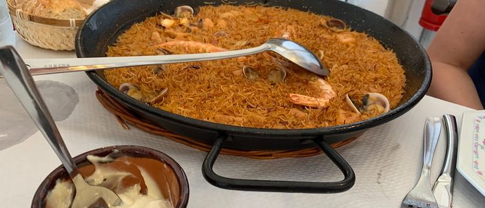 A fideuà in its wide pan on a restaurant table, prawns and clams over toasted noodles, with a clay pot of allioli in the foreground.
