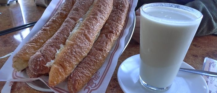 A tall glass of milky horchata beside a paper-lined plate of long glazed fartons, on a marble café counter.