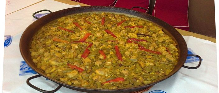 A wide steel paella pan of paella valenciana — green beans, chicken and strips of red pepper over a thin layer of rice — resting on a papered table.