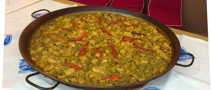 A wide steel paella pan of paella valenciana — green beans, chicken and strips of red pepper over a thin layer of rice — resting on a papered table.