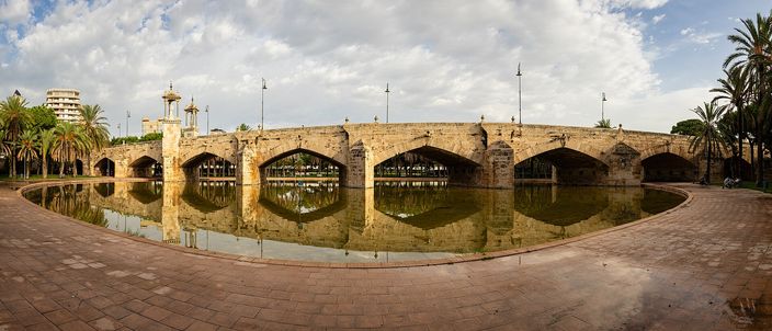 The stone arches of the Pont de la Mar reflected in a still pool in the Turia garden, palm trees on either side under a summer sky.