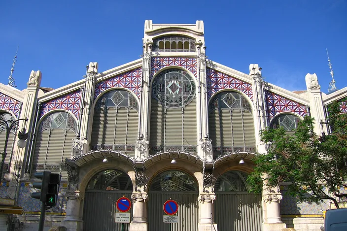 The main gable of Valencia's Mercat Central: a modernista front of stone piers and arched entrances under a huge fan of leaded colored glass, with red-and-blue tiled panels either side and a clear blue sky above.