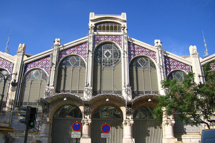The main gable of Valencia's Mercat Central: a modernista front of stone piers and arched entrances under a huge fan of leaded coloured glass, with red-and-blue tiled panels either side and a clear blue sky above.