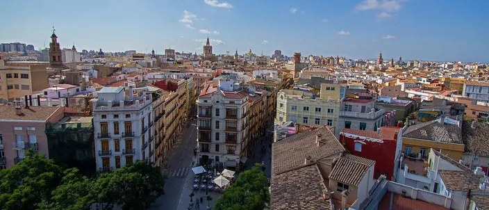 Valencia's old town seen from above: terracotta rooftops in the foreground, a straight lane of ochre and pastel apartment blocks running away from the camera, and the cathedral's octagonal bell tower and other spires on the skyline under a blue sky.