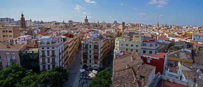 Valencia's old town seen from above: terracotta rooftops in the foreground, a straight lane of ochre and pastel apartment blocks running away from the camera, and the cathedral's octagonal bell tower and other spires on the skyline under a blue sky.
