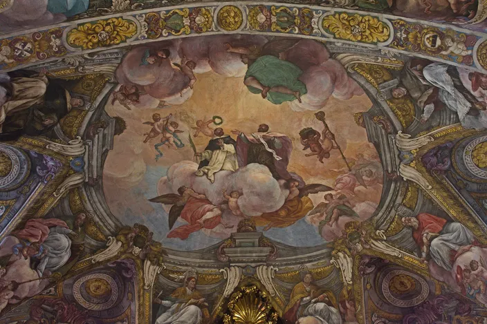 A Baroque painted church vault seen from below: cherubs and robed figures on banks of cloud filling a domed panel, framed by gilded stucco cartouches and painted saints along the ribs.