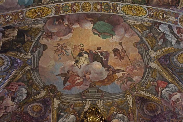 A Baroque painted church vault seen from below: cherubs and robed figures on banks of cloud filling a domed panel, framed by gilded stucco cartouches and painted saints along the ribs.