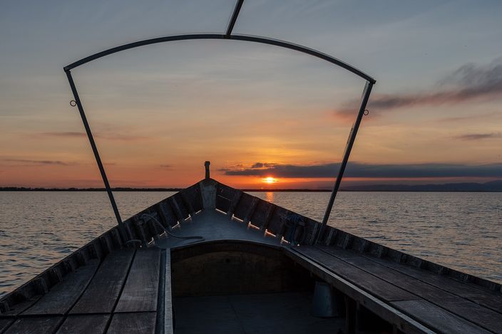 Looking forward from inside a flat wooden boat over the Albufera lagoon at sunset, the planked prow and its curved metal hoop in silhouette against an orange sky and a low sun on the water.