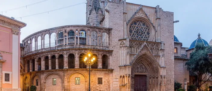 Valencia cathedral at dawn under a pale blue sky: a curved two-story arcade on the left, a tall Gothic gable with a rose window and a deeply moulded pointed doorway on the right, a lit street lamp in front of them and the square's paving wet and reflecting.