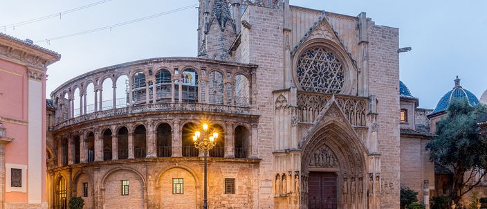 Valencia cathedral at dawn under a pale blue sky: a curved two-storey arcade on the left, a tall Gothic gable with a rose window and a deeply moulded pointed doorway on the right, a lit street lamp in front of them and the square's paving wet and reflecting.