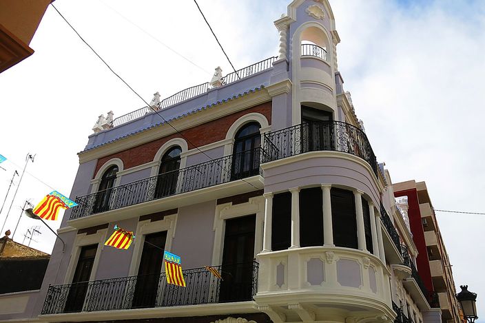 An ornate corner house in El Cabanyal with white mouldings, wrought-iron balconies and Valencian flags, its bay window rounding the street corner.