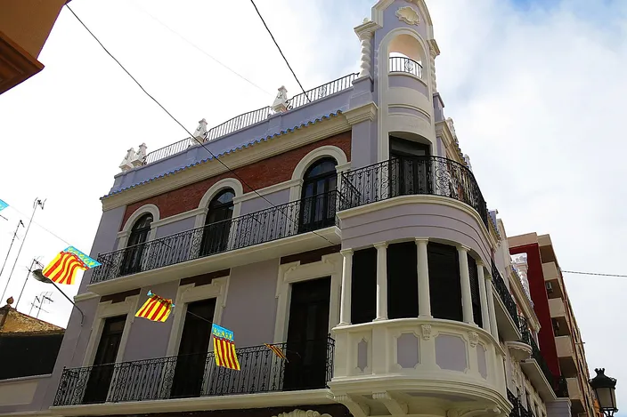 An ornate corner house in El Cabanyal with white mouldings, wrought-iron balconies and Valencian flags, its bay window rounding the street corner.