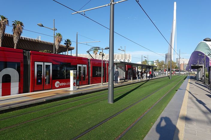 A red Metrovalencia tram on a grass-track stop, with the white cable-stay bridge and domed Àgora of the City of Arts rising behind.