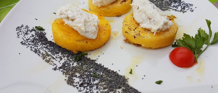 Two rounds of grilled polenta each topped with a quenelle of pale, whipped baccalà mantecato, plated with a scatter of black pepper and a cherry tomato.