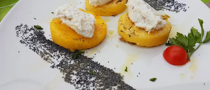 Two rounds of grilled polenta each topped with a quenelle of pale, whipped baccalà mantecato, plated with a scatter of black pepper and a cherry tomato.