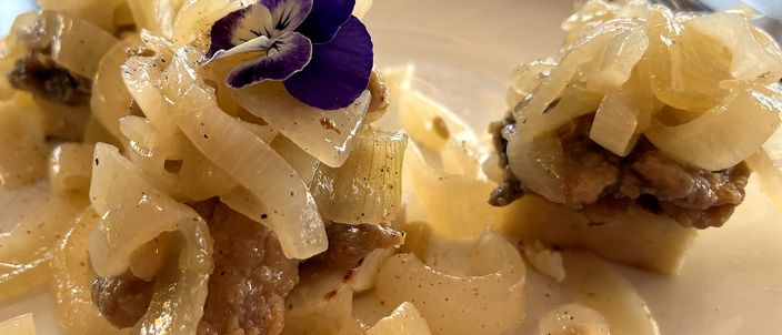 A plate of sarde in saor: fried sardines topped with a tangle of soft translucent onions and scattered raisins, garnished with a small edible flower.