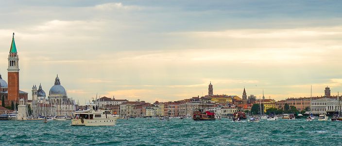 The Venice waterfront seen across the water at dusk, the green-capped campanile and domes of San Giorgio Maggiore on the left and the long line of the Riva running to the Doge's Palace on the right, boats crossing the open lagoon in front
