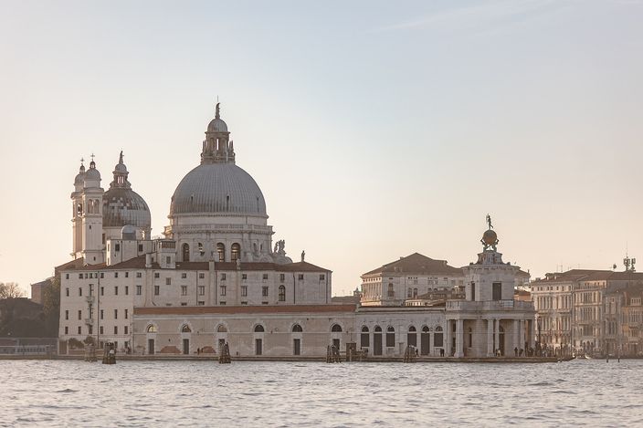 Santa Maria della Salute and the long low arcaded customs house beside it seen across open water in pale evening light, the church's great dome and two small campanili on the left, the customs house corner tower carrying a golden globe on the right, and the lagoon in the foreground