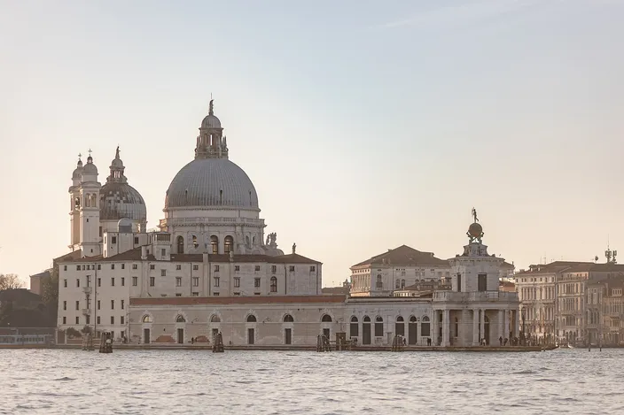 Santa Maria della Salute and the long low arcaded customs house beside it seen across open water in pale evening light, the church's great dome and two small campanili on the left, the customs house corner tower carrying a golden globe on the right, and the lagoon in the foreground