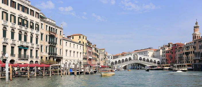 The Grand Canal seen from the water under a clear sky, palazzi with red restaurant awnings along the left bank, the white stone arch of the Rialto bridge carrying its two ranks of shops across the canal right of center, and a yellow motorboat and vaporetti crossing mid-channel
