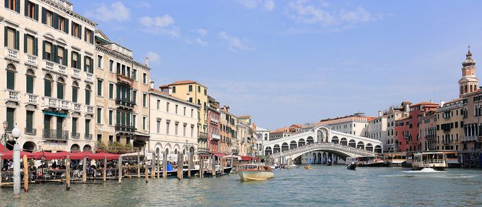 The Grand Canal seen from the water under a clear sky, palazzi with red restaurant awnings along the left bank, the white stone arch of the Rialto bridge carrying its two ranks of shops across the canal right of centre, and a yellow motorboat and vaporetti crossing mid-channel