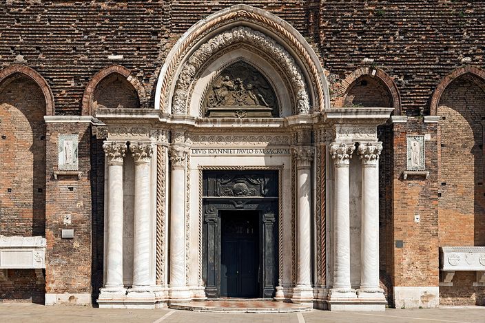 The white marble portal of Santi Giovanni e Paolo set into the church's plain brick front, a tall pointed arch carried on twisted columns with a carved relief above a dark wooden door, rows of blind brick arches running away on either side