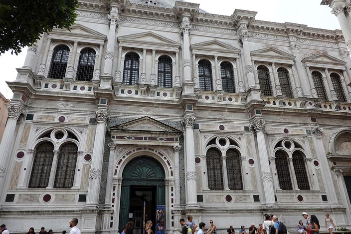 The white Istrian stone front of the Scuola Grande di San Rocco in full sun, two orders of paired columns with arched windows and coloured marble roundels between them, an open doorway in the middle, and a scatter of people sitting and standing on the paving at its foot