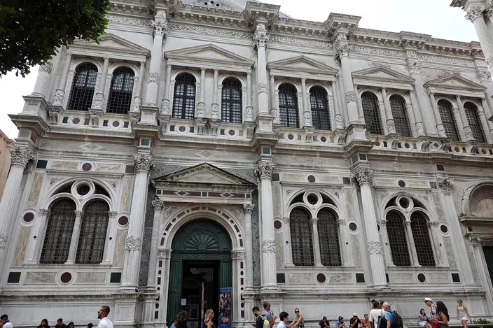 The white Istrian stone front of the Scuola Grande di San Rocco in full sun, two orders of paired columns with arched windows and colored marble roundels between them, an open doorway in the middle, and a scatter of people sitting and standing on the paving at its foot