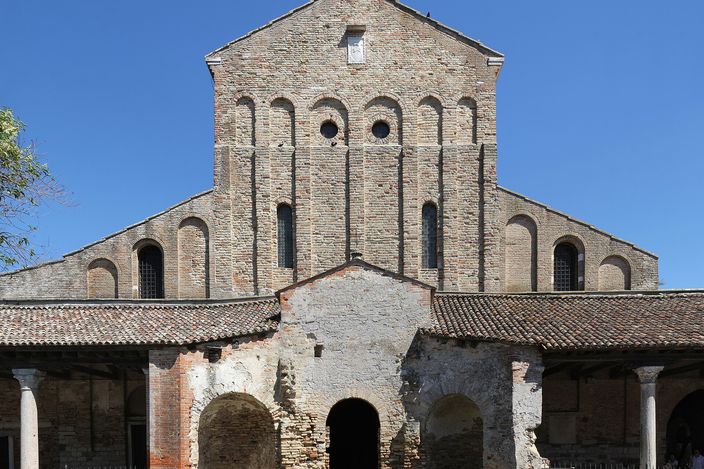 The brick front of Torcello Cathedral under a deep blue sky, a plain gabled facade with a row of blind arches, a low colonnaded portico running across its base, and the ruined arches and stone foundations of the baptistery standing in the grass in front of it