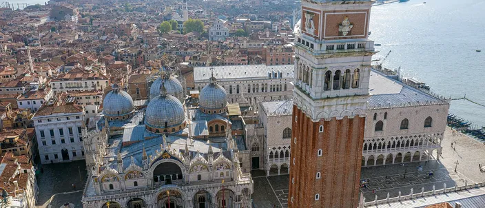 Piazza San Marco from the air in low sunlight: the five lead domes and marble front of St Mark's Basilica on the left, the brick campanile rising through the center of the frame under its green spire, the arcaded flank of the Doge's Palace and the empty piazzetta on the right, and the roofs of the city and the lagoon stretching away behind