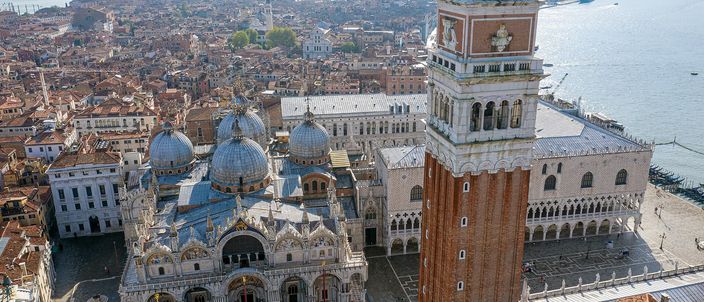Piazza San Marco from the air in low sunlight: the five lead domes and marble front of St Mark's Basilica on the left, the brick campanile rising through the centre of the frame under its green spire, the arcaded flank of the Doge's Palace and the empty piazzetta on the right, and the roofs of the city and the lagoon stretching away behind