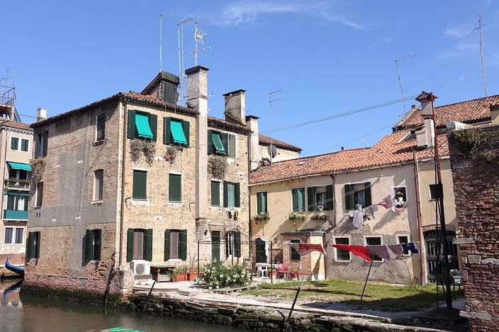 Weathered brick and stucco houses with green shutters and awnings on a small canal in Dorsoduro, washing strung across the wall to dry, plants and chairs on the paved bank, a gondola prow at the left edge