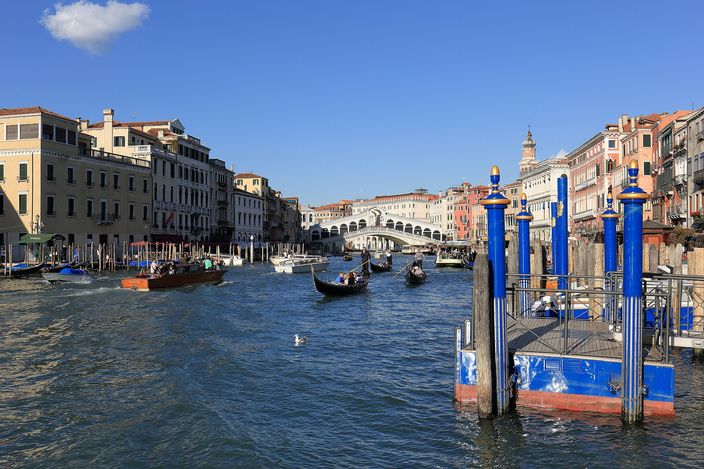 The Grand Canal busy with boats under a clear sky, looking towards the white arch of the Rialto bridge in the distance; a varnished wooden water taxi and a gondola under oar carry passengers mid-channel, more gondolas are moored along the palazzi on the left bank, and a blue-and-white vaporetto landing pontoon on tall blue mooring piles fills the right foreground
