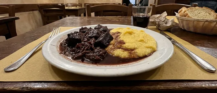 AI-generated image of pastissada de caval: dark braised meat in a glossy sauce beside soft yellow polenta on a white plate, a fork on its left and a knife on its right, in a trattoria with a glass of red wine and a bread basket behind.