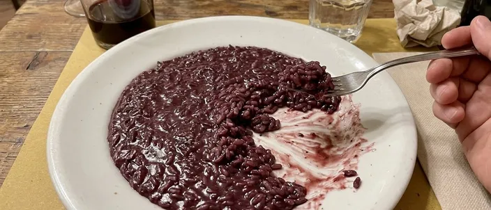 AI-generated image of risotto all'Amarone: a deep violet-red risotto spread flat across a white plate, a hand lifting some with a single fork, a glass of red wine and a glass of water behind on a trattoria table.
