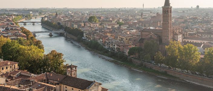 An elevated panorama of Verona's historic centre inside the bend of the Adige, with terracotta rooftops, two bridges, and a tall brick bell tower rising above the skyline in warm late-afternoon light.