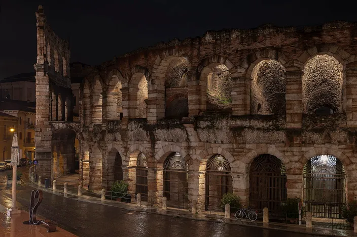 The ancient Roman arena of Verona lit up at night, its tiered stone arches glowing above a wet, empty street.