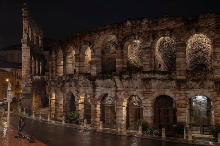 The ancient Roman arena of Verona lit up at night, its tiered stone arches glowing above a wet, empty street.
