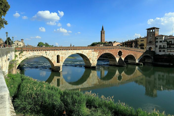 Ponte Pietra's brick and stone arches crossing the Adige in bright daylight, reflected in the calm water, a bell tower and the wooded hillside of Castel San Pietro visible beyond.