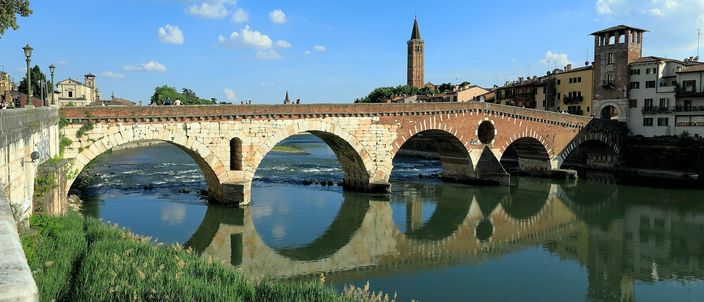 Ponte Pietra, a stone arched bridge over the Adige river in Verona, with a bell tower behind it, reflected in the calm water under a blue sky.