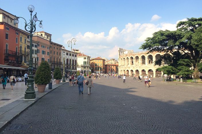 Piazza Bra in daylight, café tables and a lamppost in the foreground, the Roman Arena's stone arches filling the square's far side, tourists walking across the cobbles.