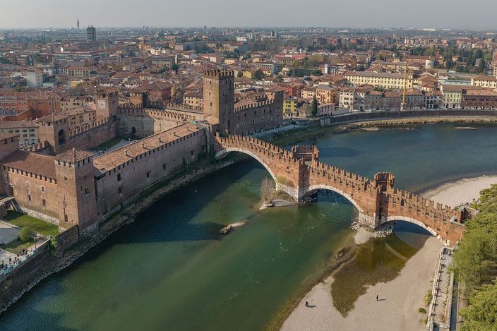 Aerial view of Castelvecchio's red-brick fortress and battlements on the Adige, with Ponte Scaligero's fortified arches crossing the river beside it.