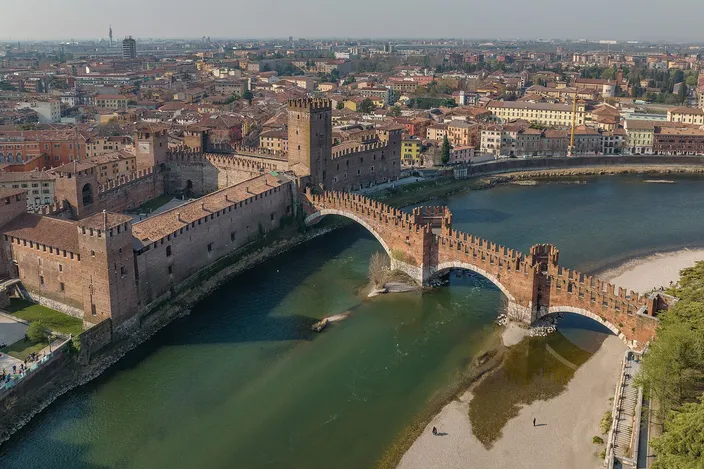 Aerial view of Castelvecchio's red-brick fortress and battlements on the Adige, with Ponte Scaligero's fortified arches crossing the river beside it.