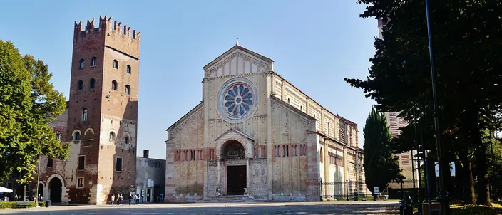 San Zeno Maggiore's Romanesque facade and campanile under a clear blue sky, the rose window and bronze-doored portal facing an empty square.