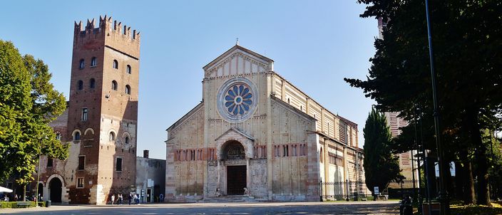 San Zeno Maggiore's Romanesque facade and campanile under a clear blue sky, the rose window and bronze-doored portal facing an empty square.